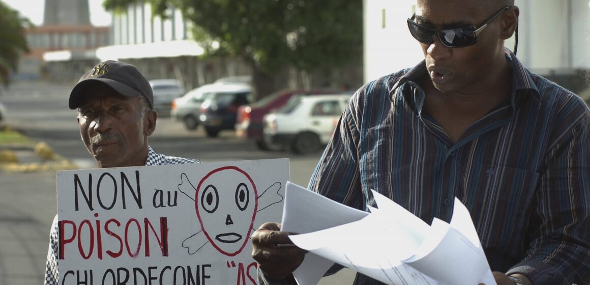 Sur cette photo du 18 novembre 2007, l'avocat Harry Durimel, à droite, se tient à côté d'un militant avec une pancarte indiquant "Non au poison du chlordécone", alors qu'ils protestent lors de l'arrivée de la ministre française de la Santé, Roselyne Bachelot à Raizet Abymes. Durimel fait alors partie des avocats représentant les travailleurs agricoles de Guadeloupe et de Martinique qui luttent depuis longtemps pour obtenir une compensation pour l'utilisation d'un pesticide interdit, le chlordécone. Photo Sipa / AP / Dominique Chomereau-Lamotte Sur cette photo du 18 novembre 2007, l'avocat Harry Durimel, à droite, se tient à côté d'un militant avec une pancarte indiquant "Non au poison du chlordécone", alors qu'ils protestent lors de l'arrivée de la ministre française de la Santé, Roselyne Bachelot à Raizet Abymes. Durimel fait alors partie des avocats représentant les travailleurs agricoles de Guadeloupe et de Martinique qui luttent depuis longtemps pour obtenir une compensation pour l'utilisation d'un pesticide interdit, le chlordécone. Photo Sipa / AP / Dominique Chomereau-Lamotte